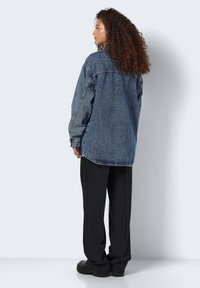 Giacca di jeans blu oversize con taglio classico, dotata di colletto a punta, maniche lunghe e dettagli cuciti. Abbinata a pantaloni neri.