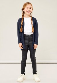 Marineblå cardigan med åpen front og lommer. Hvit t-skjorte under, svarte leggings og hvite sneakers. Jevnt stoff og tettsittende design.