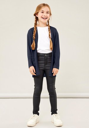 Cardigan navy avec une ouverture sur le devant, poches. T-shirt blanc en dessous, leggings noirs et baskets blanches. Tissu lisse et design ajusté.