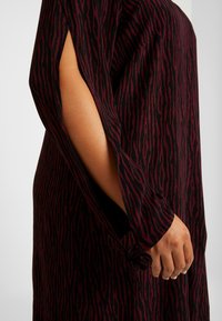 Schwarze und burgunderrote Tunika mit einer strukturierten, gestreiften Musterung. Die Ärmel haben Schlitze mit Bändern am Handgelenk für eine verstellbare Passform.