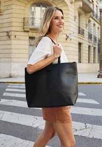 Zwarte leren tote bag met een matte afwerking, voorzien van een gestructureerde vorm, dubbele handvatten en een klein logo-detail aan de voorkant.