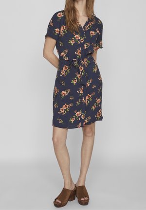 Kvinde iført en mørkeblå blomsterprintet kjole med korte ærmer, taljebånd og brune åbne sandaler med hæl, stående foran en ensfarvet baggrund.