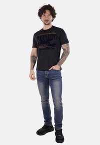 Camiseta negra con el texto "Tornado" y un gráfico de avión, combinada con jeans azules y zapatillas negras. El modelo tiene el pelo rizado y tatuajes.