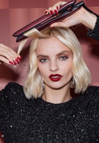 Prostownica do włosów Ghd w błyszczącej czerni z purpurowymi akcentami, trzymana nad falującymi blond włosami. Modelka ma na sobie błyszczący czarny top i czerwone paznokcie.