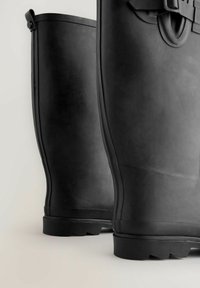 Bottes en caoutchouc noires avec une texture lisse, une forme arrondie et une semelle légèrement surélevée. Présente une boucle sur le côté et des détails décoratifs près du haut.