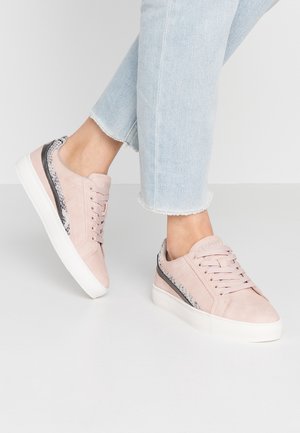 Person trägt hellrosa Low-Top-Sneaker mit weißen Sohlen und Schlangenmuster-Details, kombiniert mit hellblauen Cropped-Jeans.