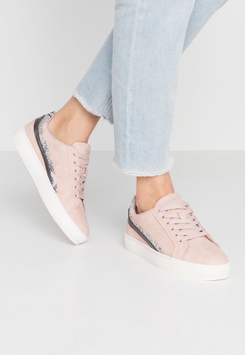 Personne portant des baskets basses rose clair avec semelles blanches et détails motif serpent, associées à un jean court bleu clair.