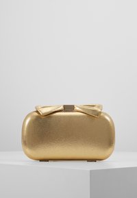 Glittrande guld rektangulär clutch med rundade kanter, prydd med en matchande tygslinga och ett metallklicklås på toppen, uppställd på en vit pelare.