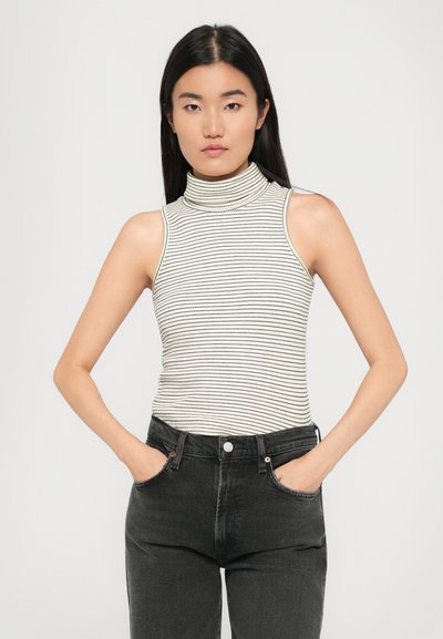 Vrouw met lang zwart haar, gekleed in een mouwloze gestreepte top met hoge nek en zwarte spijkerbroek, staand met haar handen in de zakken tegen een witte achtergrond.