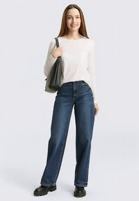 Haut blanc à manches longues associé à un jean taille haute bleu foncé et des bottes noires épaisses. Un sac fourre-tout gris est porté sur son épaule.