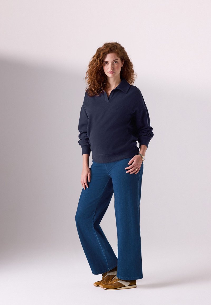 Donna con capelli rossi ricci che indossa un maglione blu navy, jeans larghi blu e sneakers marroni, in piedi davanti a uno sfondo chiaro e uniforme.