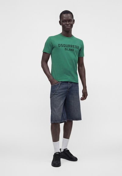 Άνδρας που φοράει πράσινο t-shirt Dsquared2 Milano, τζιν σορτς μέχρι το γόνατο με αλυσίδα, λευκές κάλτσες και μαύρα αθλητικά παπούτσια, στέκεται με το ένα χέρι στην τσέπη.