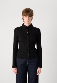 Zwarte geribbelde lange mouwen shirt met een button-down kraag en gouden knopen die langs het midden lopen. Draagt met donkere denim broek.