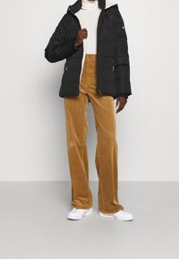 Veste noire matelassée avec capuche, associée à un pantalon large en velours côtelé beige et des baskets blanches ; textures lisses et contraste de couleurs distinct.
