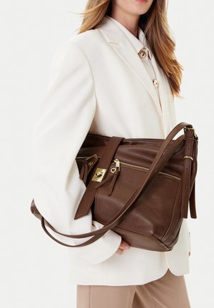 Frau in beigefarbenem Blazer, die eine große braune Lederhandtasche mit goldenen Reißverschlüssen und Schnallendetails hält.
