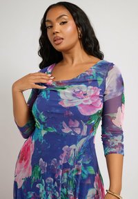 Kukkaolmuke kolmannelta neljännekseltä, sinisessä värissä, jossa on suuria vaaleanpunaisia ja vihreitä kukkakuoseja sekä laskostettu pääntie. Silkkinen ja kevyt kangas.