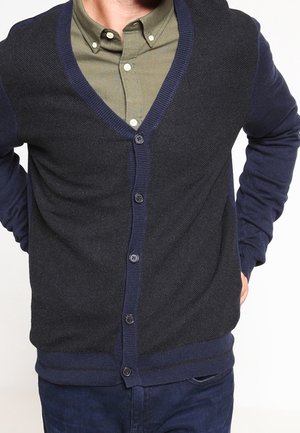 Marineblauer Cardigan mit V-Ausschnitt, durchgehender Knopfleiste, strukturiertem Strick und gerippten Bündchen und Saum. Über einem grünen Hemd mit Kragen getragen.