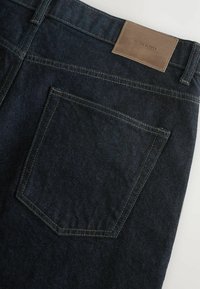 Ciemnoniebieskie dżinsy o gładkiej fakturze, z tylną kieszenią i brązową skórzaną metką z wytłoczonym napisem "DENIM SUPPLY".