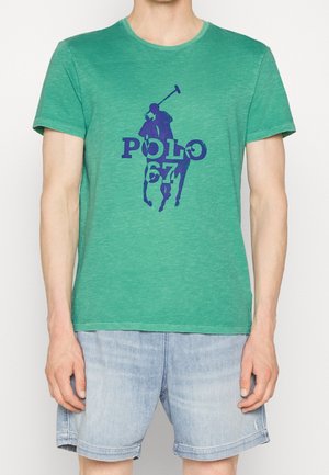 Osoba ubrana w zieloną koszulkę z krótkim rękawem z grafiką niebieskiego gracza polo i napisem "POLO 67", w połączeniu z jasnoniebieskimi szortami dżinsowymi.