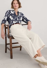 Jūras spēku un baltas krāsas rakstainā krekla ar sarullētām piedurknēm kombinācija ar platajiem bēša biksēm un gaišajām kedām, sēžot uz koka krēsla.