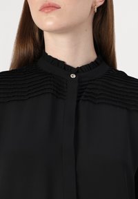 Zwarte blouse met een geplooid ontwerp langs de schouders, een hoge geschulpte kraag en een kleine decoratieve knoop bij de hals.
