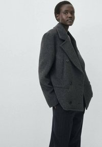 Cappotto doppio petto in lana grigia scura con rever ampi, tasche frontali e una vestibilità comoda, abbinato a pantaloni a righe.