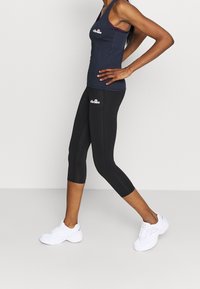 Mörkblå tanktop med en figurnära design, i kombination med svarta caprileggings. Har en logotyp på båda plaggen. Bärs med vita träningsskor.