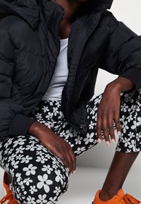 Černá bundy do pána s kapucí, žebrované manžety, spárované s černými legínami s bílými květinovými vzory, dotvořené oranžovými teniskami.