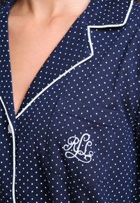 Mørkeblå pysjamas-overdel med hvite prikker, skjortekrage, kontrasterende hvite detaljer, og en frontlomme med broderte initialer.