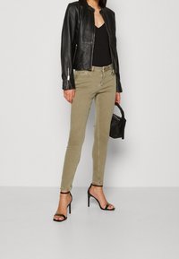 Chaqueta de cuero negra con cuello alto, jeans ajustados verde olivo y sandalias negras de tacón alto. Un bolso simple complementa el conjunto.