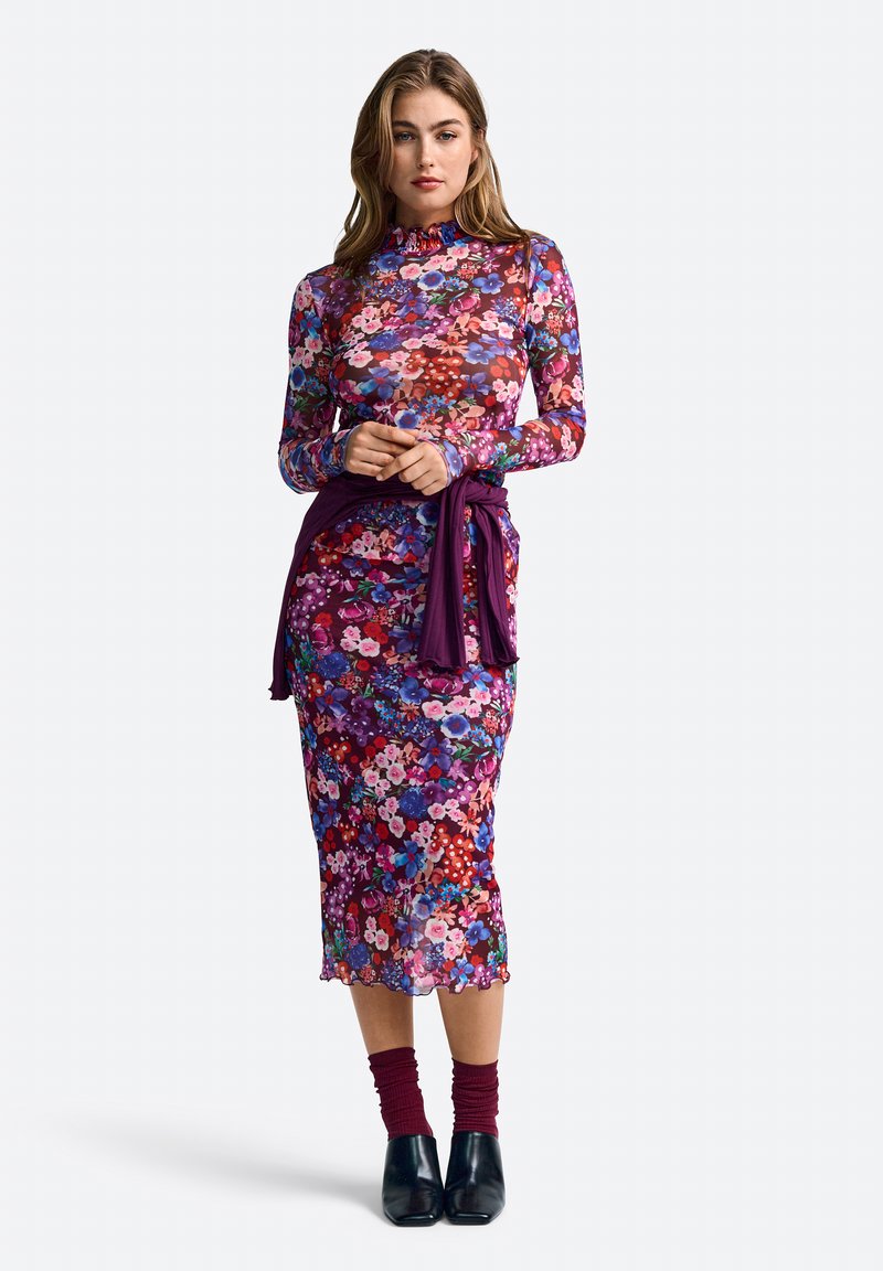 Rochie lungă cu mânecă, cu un model floral în purpură vibrantă și culori multicolore, având un guler volănaș și un accent legat la talie. Botine negre până la glezne.