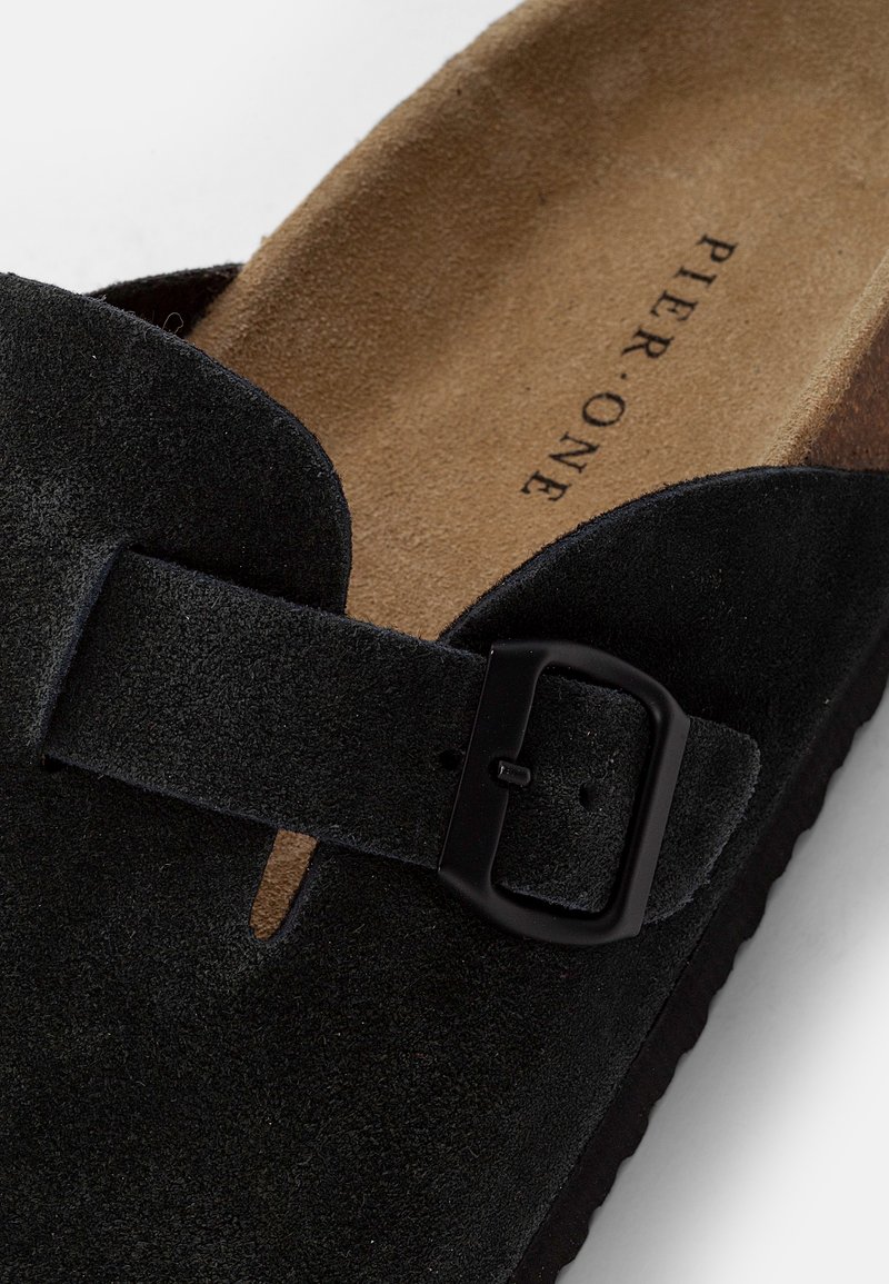 Sandale à glissière noire en suede avec une semelle synthétique, comportant une large sangle, un détail de boucle et une semelle intérieure texturée. Marque indiquée sur la semelle.
