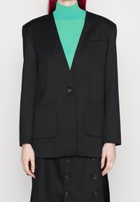Černý oversized blazer se strukturovanými rameny, zapínáním na jeden knoflík a velkými předními kapsami, doplněný tyrkysovým žebrovaným rolákem.
