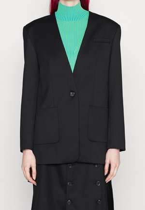 Blazer noir oversize avec des épaules structurées, fermeture à un bouton et grandes poches avant, associé à un col roulé côtelé teal.