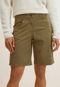 Olijfgroene shorts van lichtgewicht stof, met een ritssluiting, zakken aan de voorkant en een ontspannen pasvorm. Gestructureerde witte langemouwen top zichtbaar.