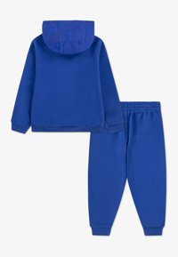 Kék pulóver és nadrág szett. A pulóvernek mintás kék kapucnija van, hosszú ujjai, és bordázott mandzsettái. A nadrág elasztikus derekú és szűkülő lábszárú.