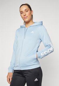 Svetlomodrá mikina na zips s kapucňou na šnúrku, s bielym logom "adidas" na ľavej hrudi a pozdĺž pravej rukávy.