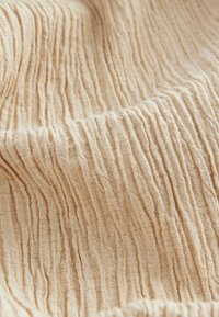 Gedeukt beige stof met een gestructureerd, gekreukt oppervlak. Het materiaal lijkt lichtgewicht en zacht met onregelmatige, lineaire patronen.