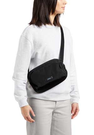 Sac bandoulière noir en tissu lisse, de forme rectangulaire, avec fermeture éclair et un patch de marque. Sangle réglable pour un port facile.