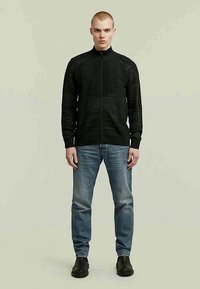 Sweater preto com fecho éclair e design de malha texturizada, combinado com jeans azul claro e botas de tornozelo pretas, em pé contra um fundo neutro.