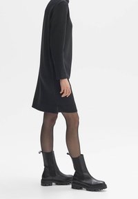 Femme portant une robe noire texturée à manches longues, des collants noirs transparents et des bottines noires épaisses, debout devant un fond blanc.