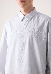 Šviesiai mėlyna dryžuota mygtukų marškiniai su balta siuvinėta "CALVIN KLEIN" ant krūtinės, turintys standartinį apykaklę ir baltas mygtukus.