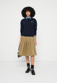 Tengerészkék pulóver kék gallérral, bézs pliszírozott szoknya, fekete cipők és csíkos zoknik. A modell egy fekete mintás táskát tart a kezében.