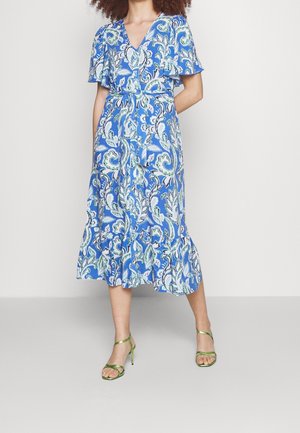 Woman wearing a blue paisley midi dress with flutter sleeves and green strappy heeled sandals, standing against a plain white background.