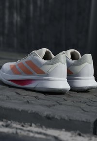 Chaussures de sport blanches avec des rayures orange et des accents roses sur une surface en pierre texturée, photographiées par l'arrière.