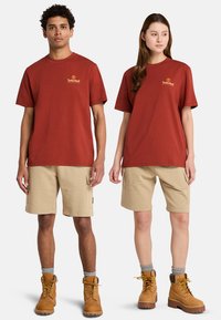 Camisetas de algodón rojas con el logo de Timberland, combinadas con pantalones cortos de cargo beige. Ambos modelos llevan botas marrones con cordones amarillos y calcetines grises.