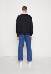 Hombre con sudadera negra, jeans anchos de pierna recta y puños azules, calcetines blancos y zapatillas blancas, de pie frente a una pared gris clara simple.