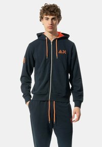Felpa con zip di colore navy con cordini arancioni, logo sul petto e pantaloni coordinati. Realizzata in tessuto morbido con polsini e vita a coste.
