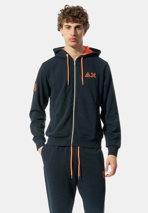 Felpa con zip di colore navy con cordini arancioni, logo sul petto e pantaloni coordinati. Realizzata in tessuto morbido con polsini e vita a coste.
