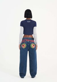 Maglietta a maniche corte blu navy sovrapposta a una maglietta a maniche lunghe di colore grigio chiaro, abbinata a jeans azzurri larghi con ricami grafici colorati sui taschini posteriori.